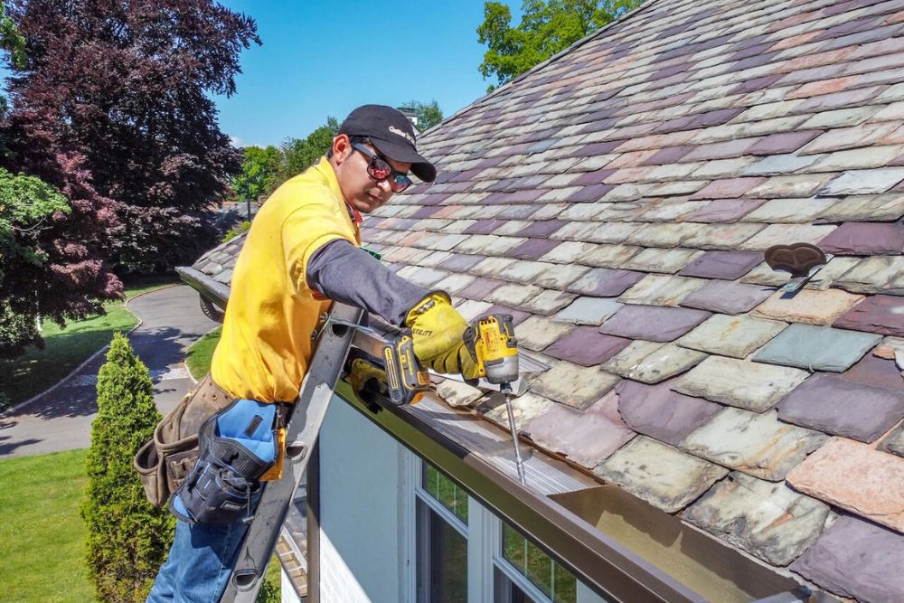 A man on a ladder working on a roof, providing gutter service to ensure proper drainage.