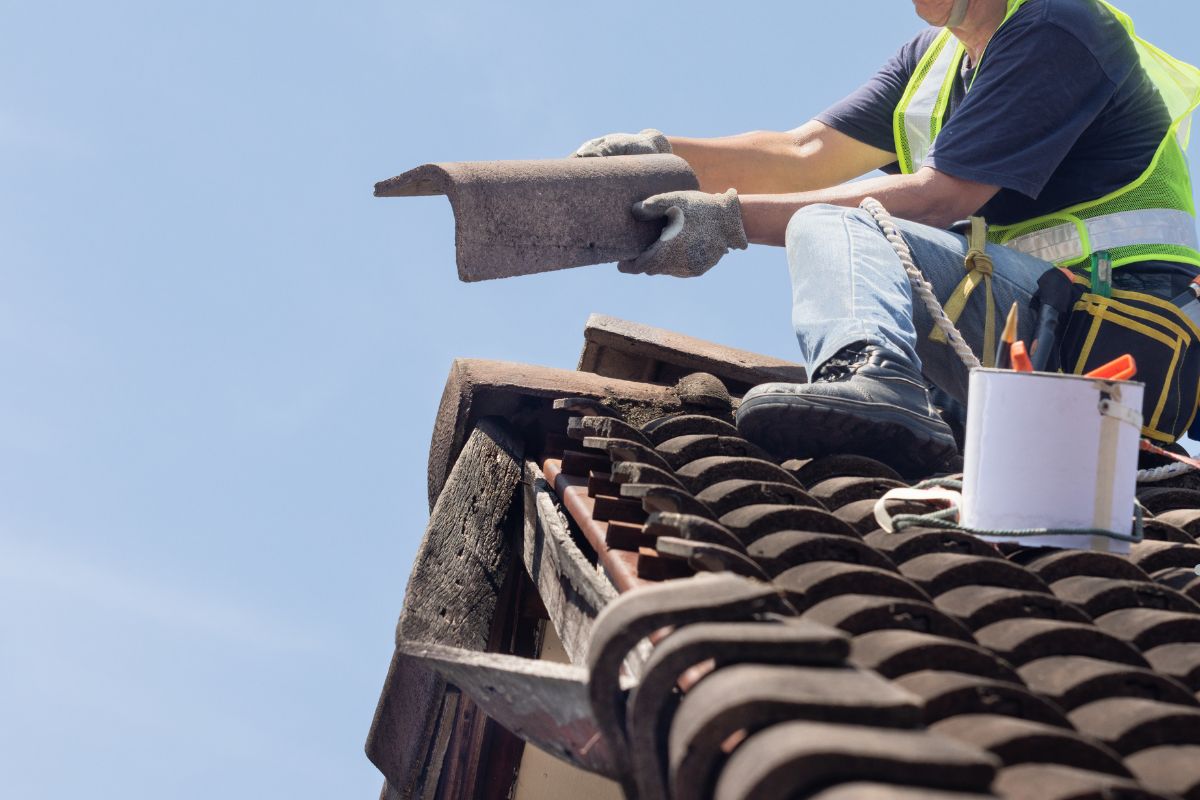 A roofer in a safety vest installs a tile on a sloped roof, with tools and a bucket nearby, against a clear blue sky.