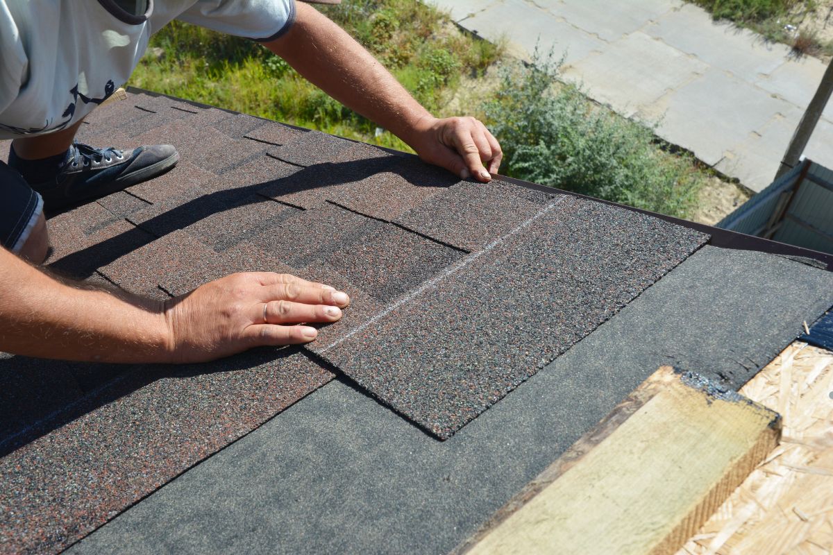 A man installs a roof shingle on a house, using tools and standing on a ladder for support.