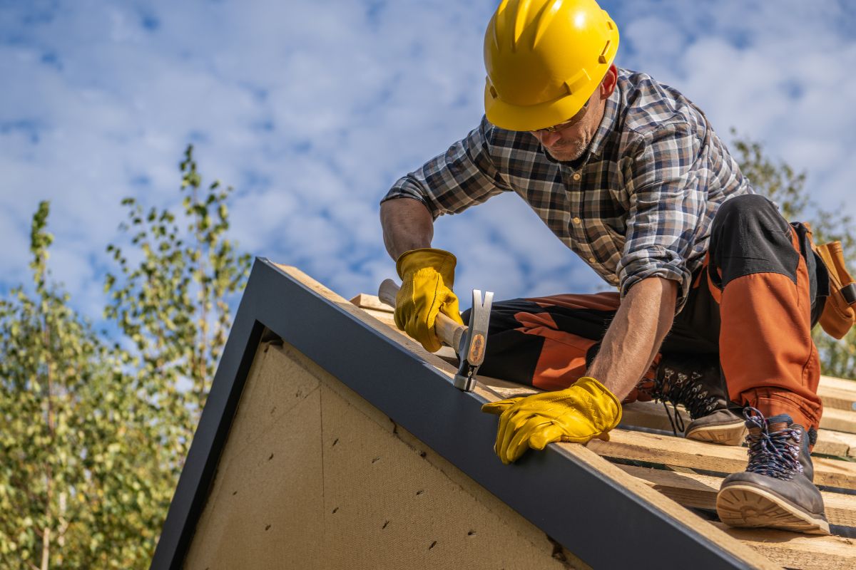 A construction worker in a yellow hard hat and gloves uses a hammer on a wooden roof structure under a blue sky.