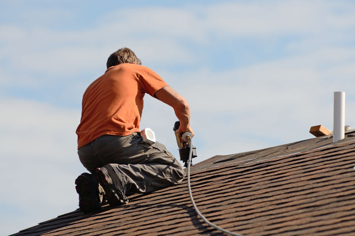 A worker in an orange shirt uses a nail gun while sitting on a sloped roof under a blue sky with scattered clouds.