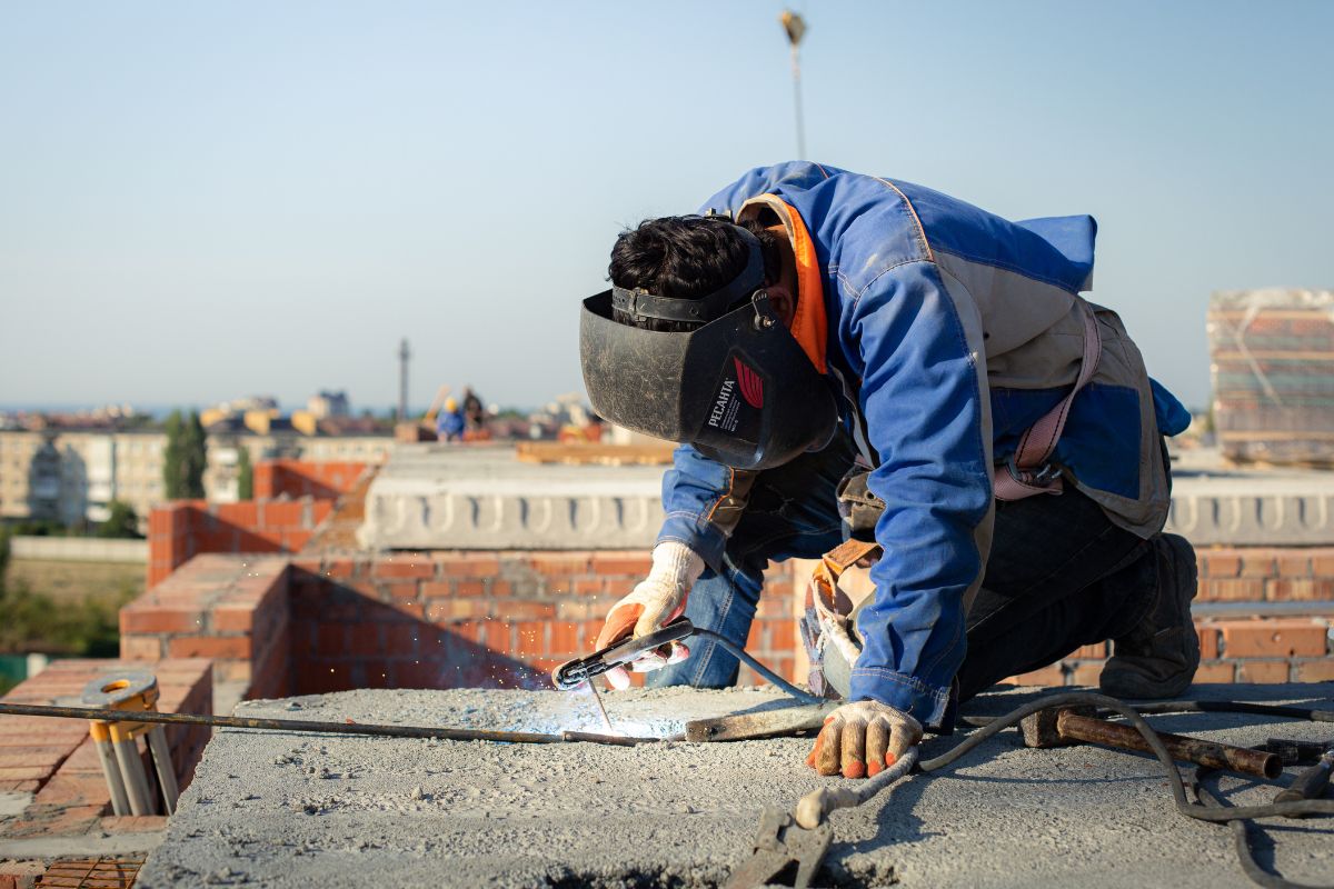 A construction worker in protective gear welds on a rooftop, with sparks flying against a clear blue sky and a cityscape in the background.