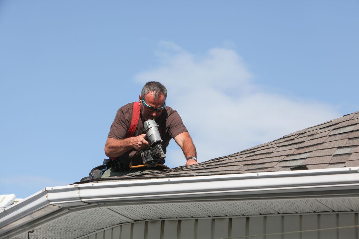 A person on a roof, using a nail gun to install shingles, set against a clear blue sky.